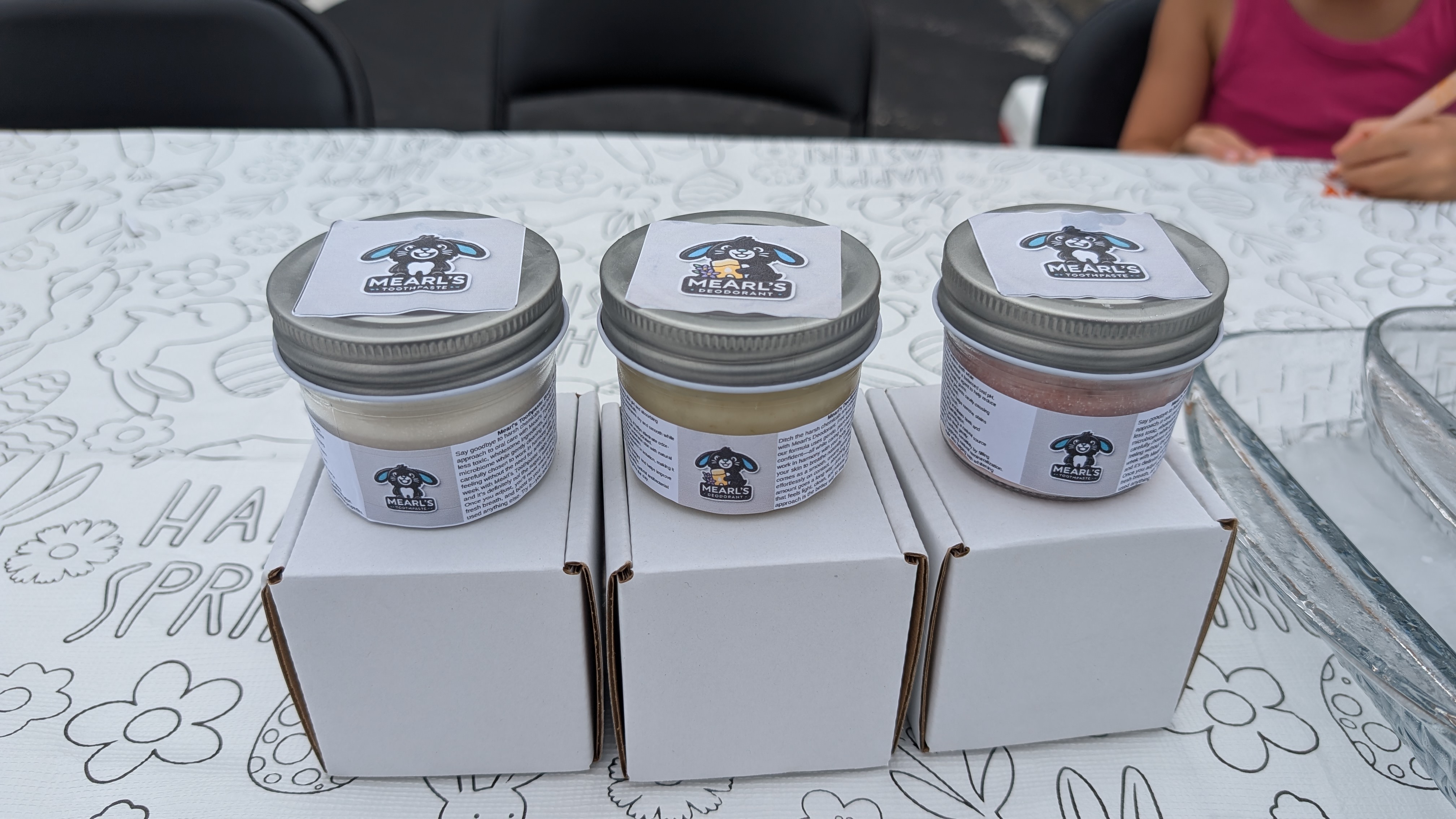 Three clear glass Mearl’s product jars with silver-colored metal lids displayed at a market table.